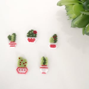 May include: Five small beaded cactus plants in pots. The cacti are green with red accents and the pots are white with pink accents. The cacti are arranged on a white surface.