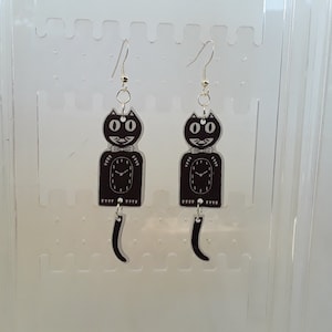 May include: A pair of black cat clock earrings with silver clock hands and a dangling tail. The earrings are hanging from silver hooks.