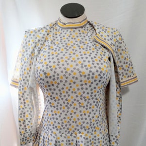 May include: A vintage white dress and matching jacket set. The dress and jacket are covered in a pattern of yellow and gray clover-like shapes. The dress has a pleated skirt and a high neckline. The jacket has short sleeves.