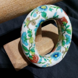 May include: A white ceramic bangle with a floral design in shades of blue, green, red, and yellow. The bangle is decorated with a repeating pattern of flowers and leaves.