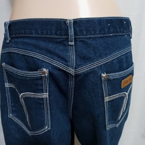 May include: Dark blue denim jeans with white stitching detail and a brown leather patch with the word "GRANDE" on the back right pocket.