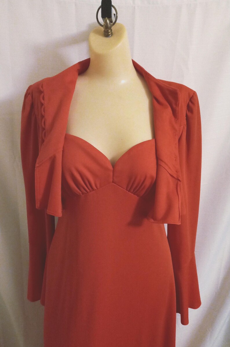 May include: A long, rust-colored dress with a matching jacket. The dress has a sweetheart neckline and long bell sleeves. The jacket has a ruffled collar and long sleeves.