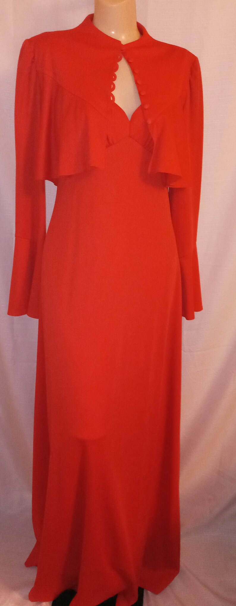 May include: A long red dress with a high neckline and a ruffled collar. The dress has a keyhole opening at the neckline and is fastened with buttons. The dress has long sleeves with bell bottoms.