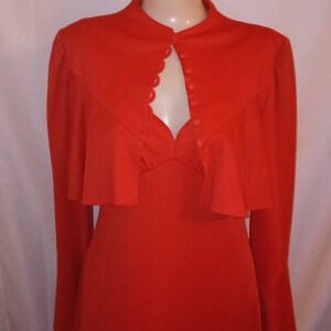 May include: A long-sleeved, red dress with a high neckline and a scalloped trim around the neckline. The dress has a button closure at the front and a ruffled overlay at the shoulders.