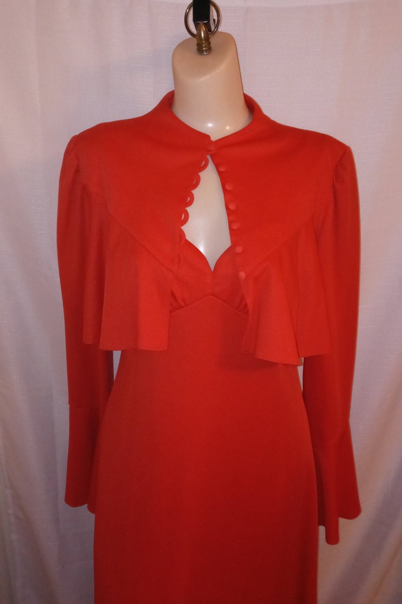 May include: A long-sleeved, red dress with a high neckline and a scalloped trim around the neckline. The dress has a button closure at the front and a ruffled overlay at the shoulders.