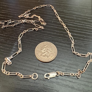 May include: A rose gold-toned paperclip chain necklace featuring elongated rectangular links and a lobster claw clasp. A United States quarter coin is placed next to the chain to provide a visual reference for the scale of the metal links.