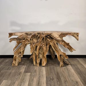 May include: A console table crafted from a single piece of light brown natural teak wood root. The table features a smooth, flat top surface supported by an intricate, organic base formed by the gnarled, interlocking branches of the tree root.