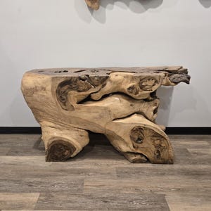 May include: A console table crafted from a single piece of natural light-colored wood with a live edge top. The base features organic, gnarled wood textures with dark brown knots and swirling grain patterns throughout the entire structure.