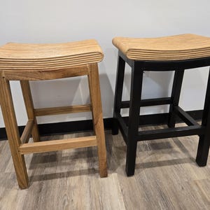 May include: Two wooden bar stools. One stool is natural wood, the other is black. Both have a curved seat and a footrest. The natural wood stool has a light brown seat and legs. The black stool has a light brown seat and black legs.