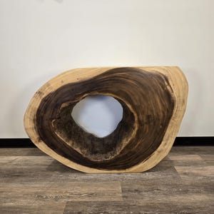 May include: A unique, live-edge wooden console table with a large, oval-shaped opening in the center. The table features a rich, dark brown grain pattern with lighter wood tones around the edges. The table is set against a white wall and a wood-look floor.