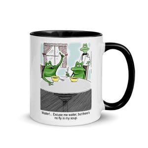 May include: White ceramic mug with a black interior and handle. The mug features a cartoon illustration of two green frogs at a table in a restaurant, with a waiter frog. Text on the mug reads: "Waiter!... Excuse me waiter, but there's no fly in my soup."