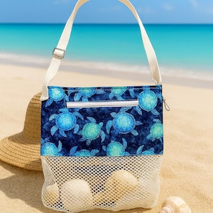 May include: A beach bag with a white mesh bottom and a blue turtle pattern top. The bag has a white strap and is filled with seashells. A straw hat sits nearby on the sand, with the ocean in the background.