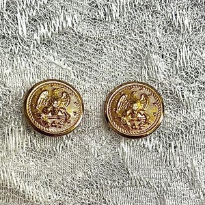 May include: Two circular gold-toned metal buttons featuring a raised relief design of an eagle perched atop an anchor, surrounded by a series of small stars along the outer rim, set against a textured white lace fabric background.