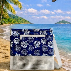 May include: A navy blue and white seashell-patterned beach bag with a white mesh bottom and a zipper closure. The bag has a white shoulder strap and is set on a sandy beach with seashells.