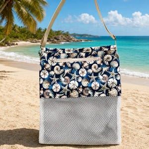 May include: A beach bag with a navy blue top section patterned with seashells and starfish, and a white mesh bottom. The bag has a beige shoulder strap and a zippered top. The background shows a sandy beach and ocean.