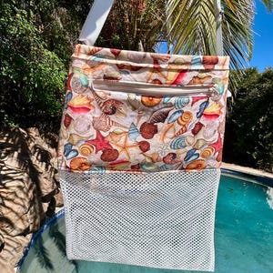 May include: A rectangular crossbody bag featuring a colorful seashell and starfish print on the upper fabric section, a horizontal zippered pocket, and a white mesh lower section. The bag includes a white adjustable shoulder strap for carrying.