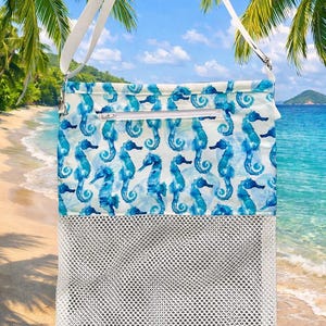 May include: A beach tote bag with a white mesh bottom and a top section featuring a blue seahorse pattern on a white background. The bag has a white shoulder strap and a zippered top closure. The background shows a beach scene with palm trees and ocean.