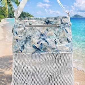 May include: A white mesh beach bag with a white strap and a top section featuring a blue and white starfish pattern. The bag has a zipper closure and is designed for carrying beach essentials.