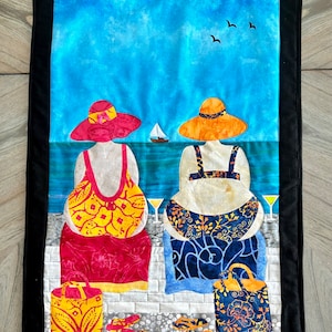 May include: Two women in colorful beachwear and hats sit on a beach with their bags and sandals. The background is a blue sky with a sailboat and birds.