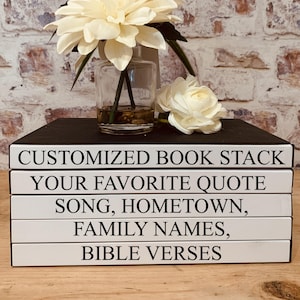 May include: A stack of white books with black text on the spines. The text reads "Customized Book Stack", "Your Favorite Quote", "Song, Hometown", "Family Names", and "Bible Verses". The books are sitting on a black surface with a white flower arrangement in a clear glass vase.