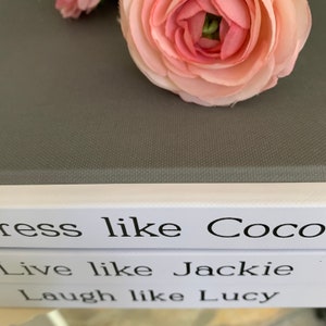 May include: Three white books stacked on top of each other. The top book has the text "Dress like Coco" printed on it. The middle book has the text "Live like Jackie" printed on it. The bottom book has the text "Laugh like Lucy" printed on it.