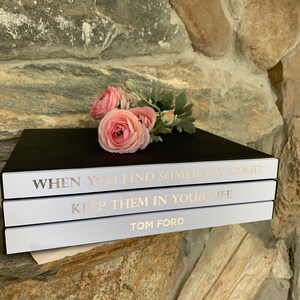 May include: Three white books stacked on top of each other. The top book has the text "WHEN YOU FIND SOMEBODY GOOD" in gold lettering. The middle book has the text "KEEP THEM IN YOUR LIFE" in gold lettering. The bottom book has the text "TOM FORD" in gold lettering. A bouquet of pink flowers is resting on the top book.