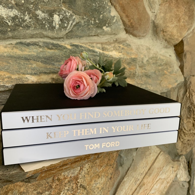 May include: Three white books stacked on top of each other. The top book has the text "WHEN YOU FIND SOMEBODY GOOD" in gold lettering. The middle book has the text "KEEP THEM IN YOUR LIFE" in gold lettering. The bottom book has the text "TOM FORD" in gold lettering. A bouquet of pink flowers is resting on the top book.