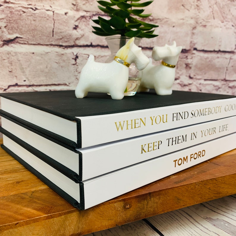 May include: Three black and white hardcover books stacked on a wooden surface. The top book has the text "WHEN YOU FIND SOMEBODY GOOD KEEP THEM IN YOUR LIFE TOM FORD" in gold lettering.