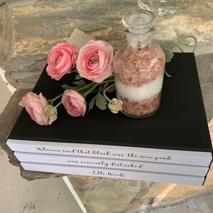 May include: Three white books stacked on top of each other with black text on the top book that reads "Whoever said that black was the new pink was seriously disturbed. - Elle Woods". A glass jar filled with pink Himalayan salt and white bath salts sits on top of the books. Pink flowers are in the background.
