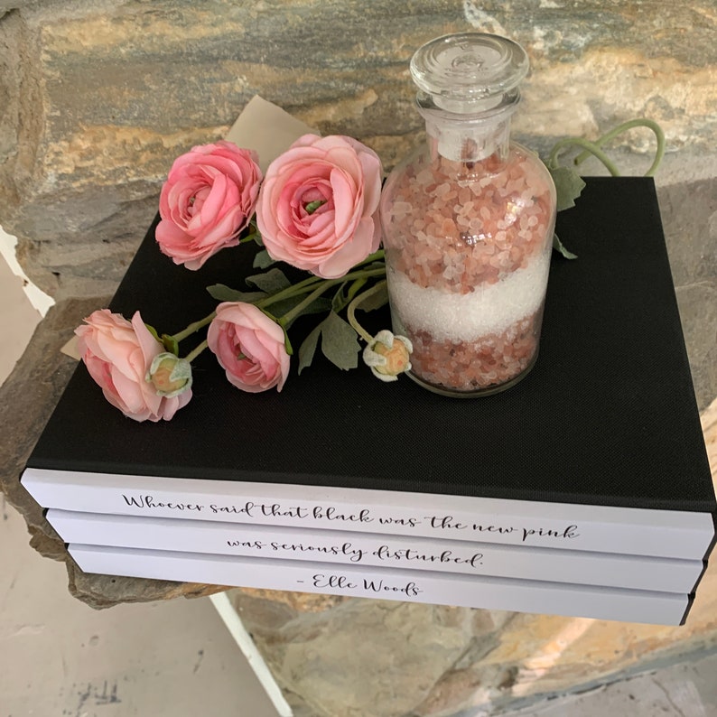 May include: Three white books stacked on top of each other with black text on the top book that reads "Whoever said that black was the new pink was seriously disturbed. - Elle Woods". A glass jar filled with pink Himalayan salt and white bath salts sits on top of the books. Pink flowers are in the background.