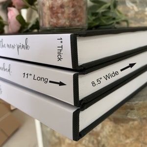 以下が含まれることがあります： 黒色の背表紙を持つ白いハードカバーの本が3冊積み重ねられています。一番上の本には「the new pink」と「1インチ厚」の文字が書かれています。真ん中の本には「11インチ長」の文字が書かれています。一番下の本には「8.5インチ幅」の文字が書かれています。