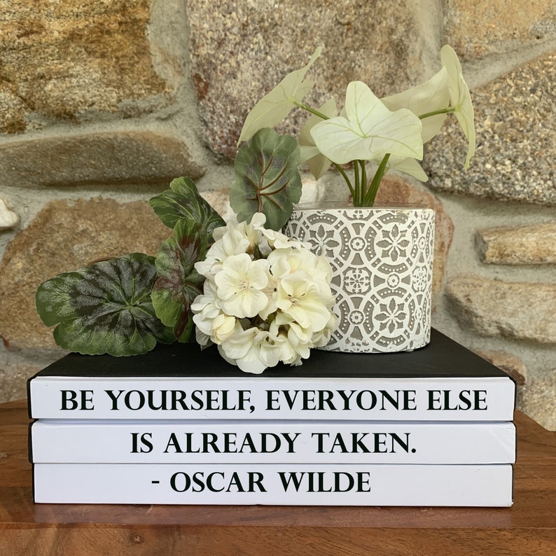 May include: A stack of three white books with black text. The top book reads "Be yourself, everyone else is already taken. - Oscar Wilde". A white ceramic planter with a grey geometric pattern holds a green plant with white flowers.