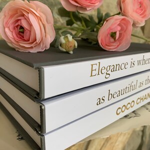 May include: A stack of three books with grey covers. The top book has white pages with the text "Elegance is when" in gold lettering. The middle book has white pages with the text "as beautiful as the" in gold lettering. The bottom book has white pages with the text "COCO CHAN" in gold lettering. The books are decorated with pink flowers.
