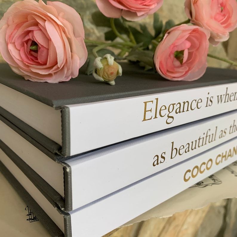May include: A stack of three books with grey covers. The top book has white pages with the text "Elegance is when" in gold lettering. The middle book has white pages with the text "as beautiful as the" in gold lettering. The bottom book has white pages with the text "COCO CHAN" in gold lettering. The books are decorated with pink flowers.
