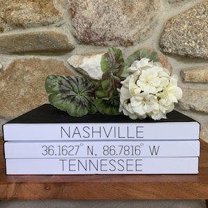 May include: Three white books stacked on top of each other. The top book is black and says "NASHVILLE" in white text. The middle book says "36.1627&deg; N, 86.7816&deg; W" in white text. The bottom book says "TENNESSEE" in white text.