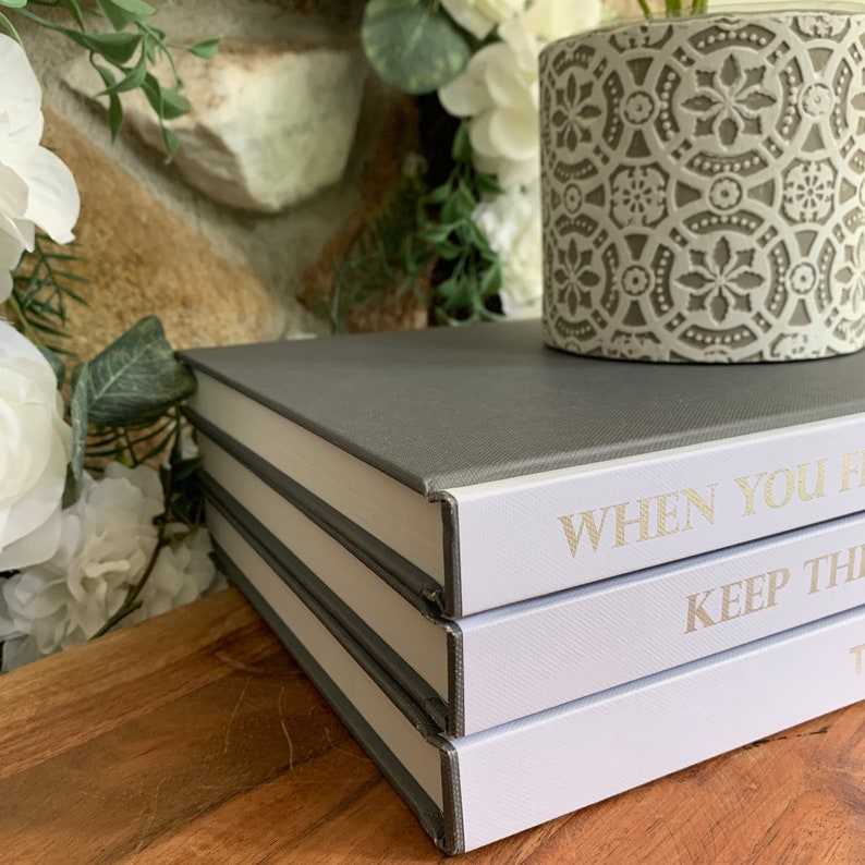 May include: Three stacked books with gray covers and white spines. The top book has the text "WHEN YOU FEEL" on the spine. The middle book has the text "KEEP THE" on the spine. The bottom book has the text "T" on the spine. The books are sitting on a wooden surface.