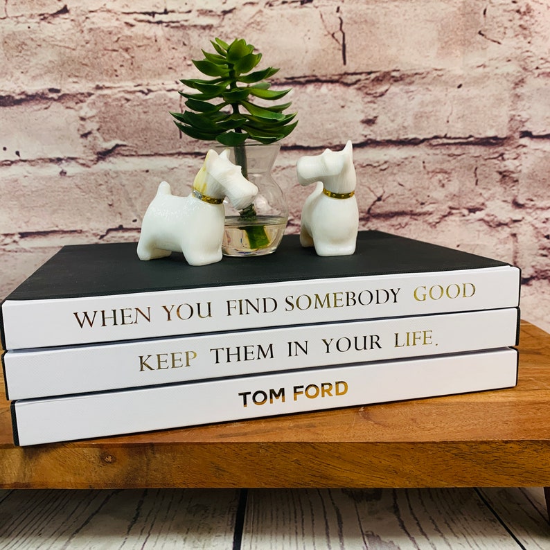 May include: Three white books stacked on a wooden surface. The top book has black cover with gold lettering that reads "WHEN YOU FIND SOMEBODY GOOD KEEP THEM IN YOUR LIFE." The middle book has a white cover with gold lettering that reads "KEEP THEM IN YOUR LIFE." The bottom book has a white cover with gold lettering that reads "TOM FORD."