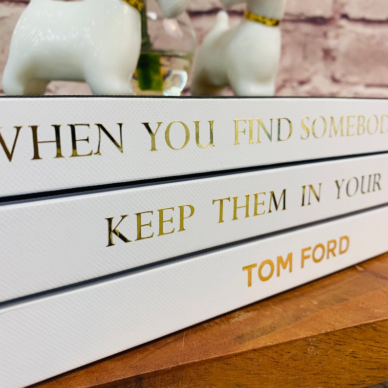 May include: Three white books stacked on top of each other. The top book has the text "WHEN YOU FIND SOMEBODY" in gold lettering. The middle book has the text "KEEP THEM IN YOUR" in gold lettering. The bottom book has the text "TOM FORD" in gold lettering.
