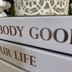 May include: Two white books stacked on top of each other. The top book has the words "BODY GOOD" printed in gold lettering. The bottom book has the words "YOUR LIFE" printed in gold lettering.