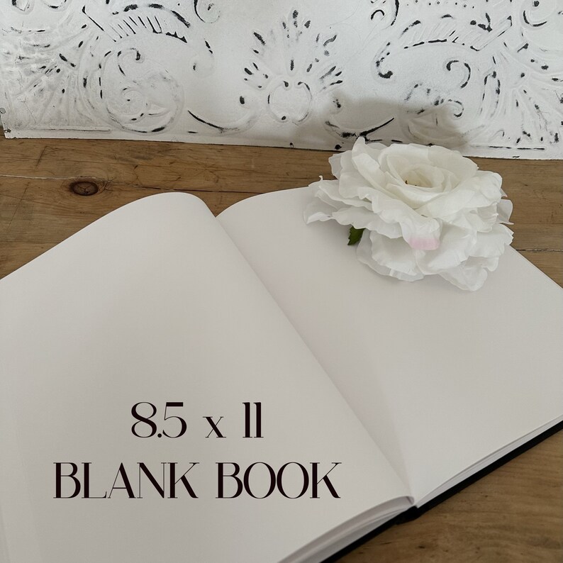 以下が含まれることがあります： 白い表紙に黒い文字で「8.5 x 11 BLANK BOOK」と書かれた空白のノート。ノートは開いて木製のテーブルの上に置かれている。ノートの右側に白い花が置かれている。