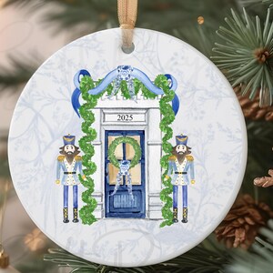 May include: Round ceramic Christmas ornament featuring a blue door with a wreath and the year 2025. Two nutcracker figures stand on either side of the door. The ornament has a gold ribbon for hanging and is decorated with a floral pattern.