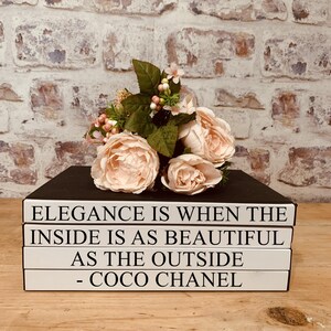 May include: A stack of four white books with black text. The top book reads "Elegance is when the inside is as beautiful as the outside - Coco Chanel". A bouquet of pink roses with green leaves sits on top of the books.