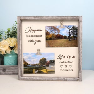 May include: A rustic wooden picture frame with two photo clips. The frame has a white background with the text "Happiness is a moment with you." and "Life is a collection of moments" printed on it. Two photos are displayed in the frame, one of a park with trees and another of a street with trees.