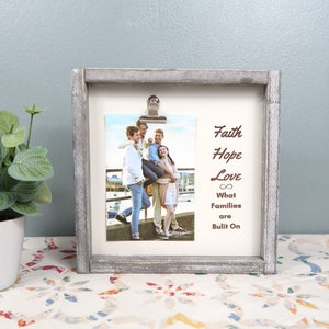 May include: A rustic gray wood picture frame with a clip holding a photo of a family of four. The frame has the words "Faith Hope Love What Families Are Built On" printed on it.