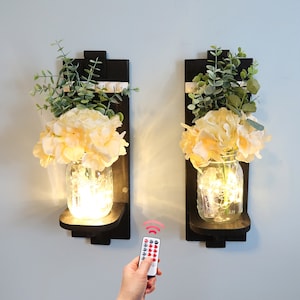 May include: Two black wall sconces with a white ribbon and a mason jar with a string of lights inside. Each sconce has a bouquet of yellow flowers and green leaves.