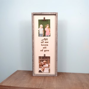 May include: A rectangular wooden photo frame with a distressed gray finish. The frame holds two photos secured by clips, with the text "All of me loves all of you" printed in brown. The photos show two people.