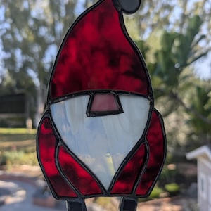 May include: A stained glass ornament in the shape of a gnome. The gnome has a red hat and coat, a white beard, and black shoes. The ornament is hanging from a silver wire.
