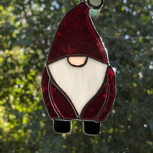 May include: A stained glass gnome ornament with a burgundy hat and coat, a white beard, and black shoes. The ornament is hanging in front of a blurred green background. The gnome has a small, round face.