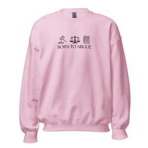 May include: A light pink sweatshirt with the text "BORN TO ARGUE" and three black icons: a gavel, scales of justice, and a law book. The sweatshirt has long sleeves and a crew neck.