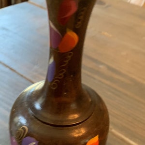 Puede incluir: Un jarrón marrón con la parte superior acampanada y una base redondeada. El jarrón está decorado con diseños florales coloridos y estilizados en tonos de rojo, morado, naranja y verde. El jarrón está sobre una mesa de madera.